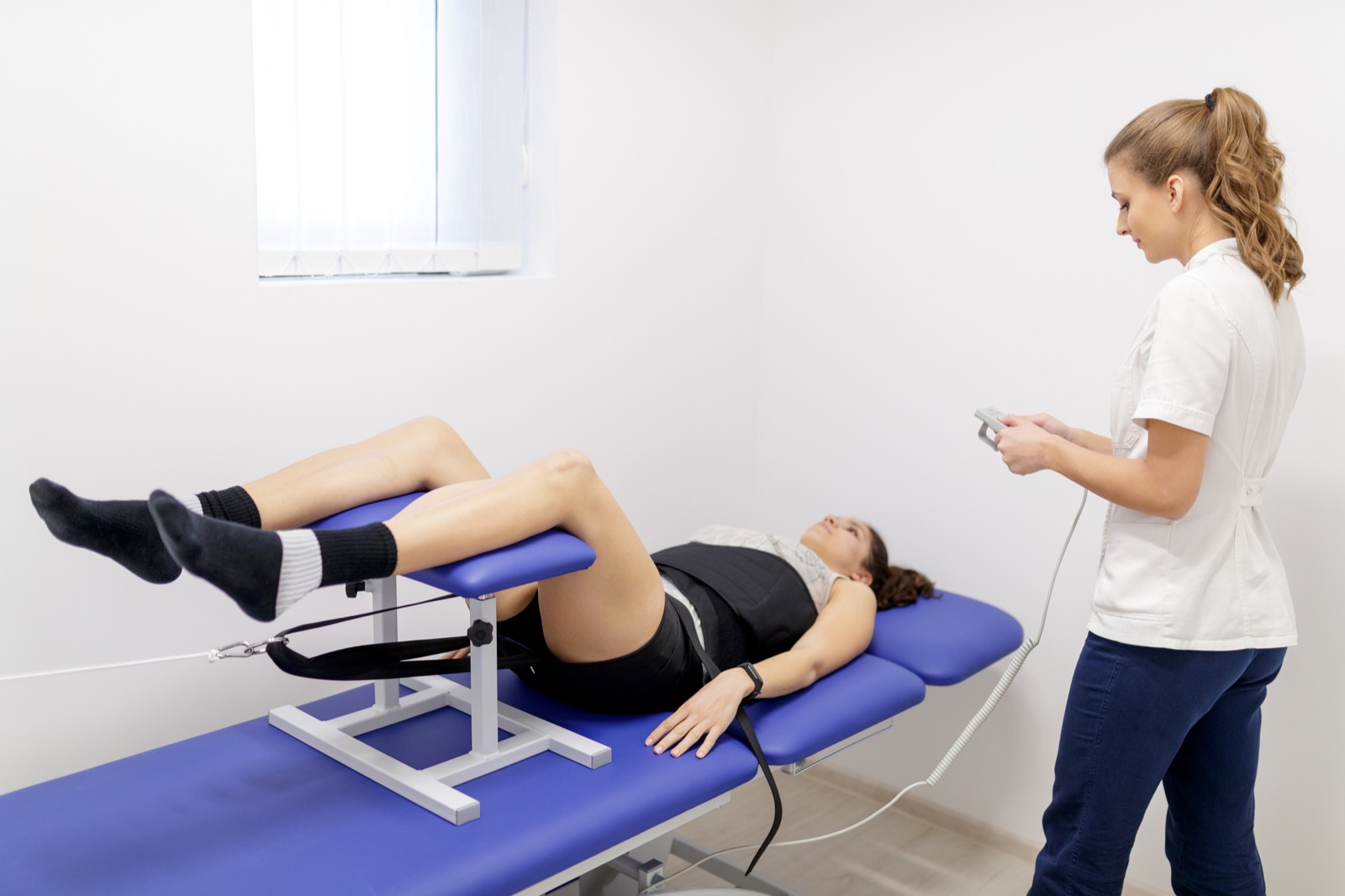 A patient on a treatment couch with their legs supported by lumbar traction equipment, while a practitioner operates the control unit